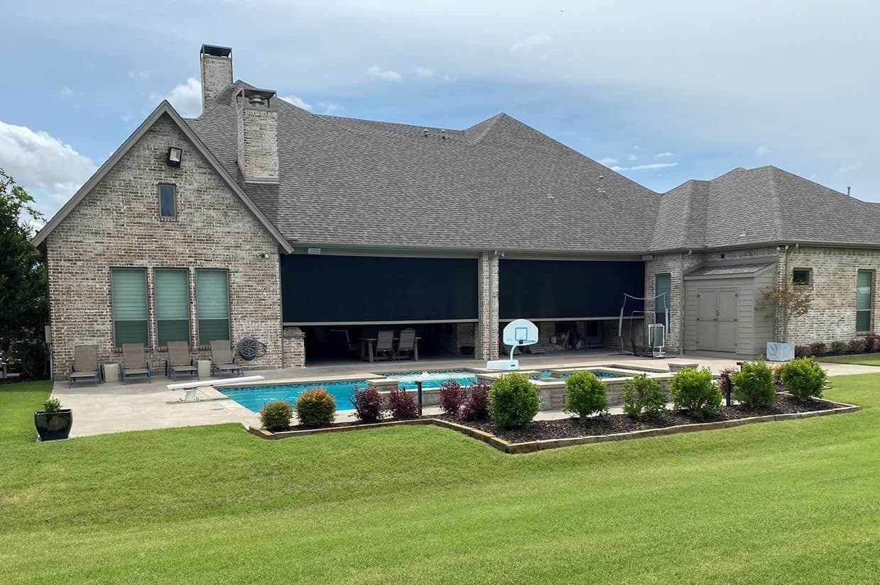 Wide exterior view of a brick house featuring dark motorized retractable privacy screens over a covered patio, facing a swimming pool and landscaped lawn.