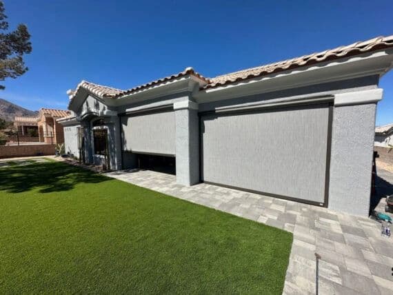 Exterior view of a stucco home with a tile roof, featuring large motorized retractable shade screens installed over the patio, set beside a paved walkway and green lawn under a clear blue sky.