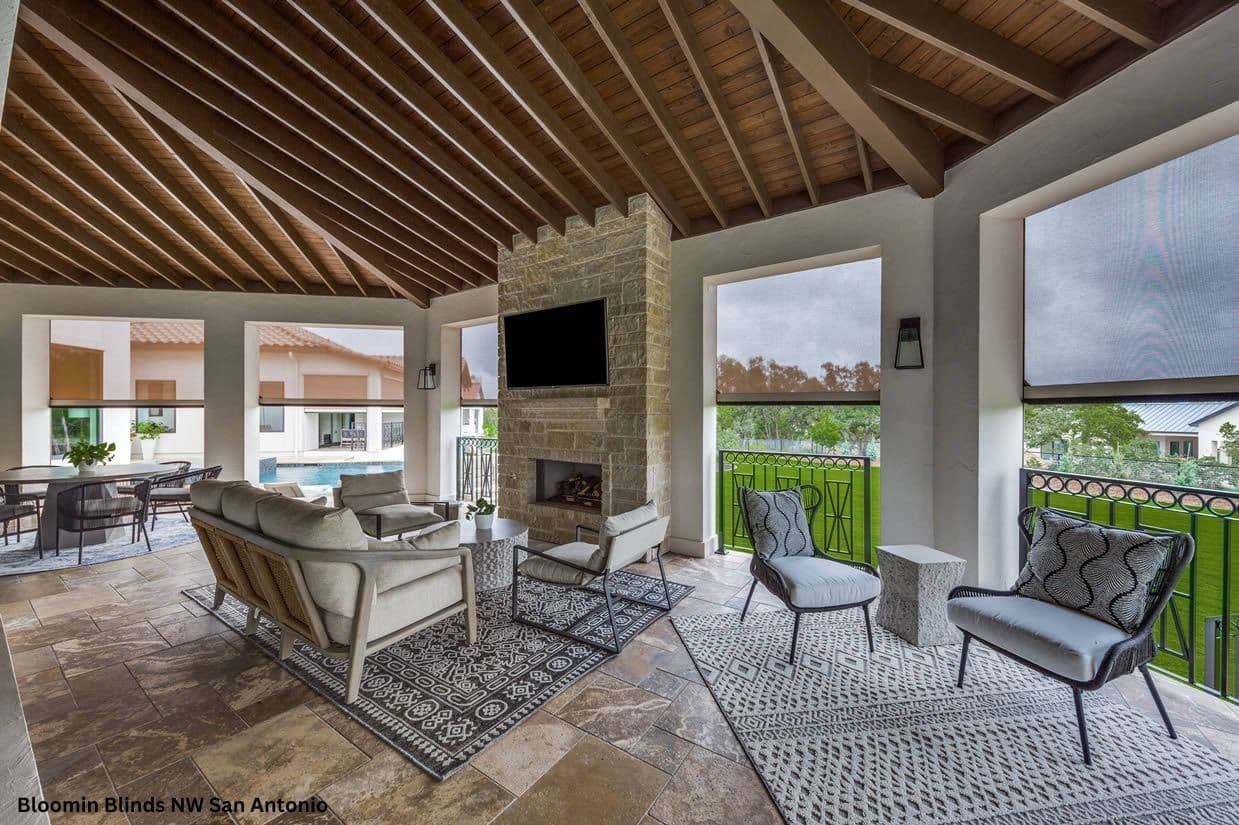 Interior view of a covered outdoor patio with exposed wood beam ceilings, stone fireplace, modern lounge seating, and retractable motorized shade screens over open archways overlooking a lawn and pool area.