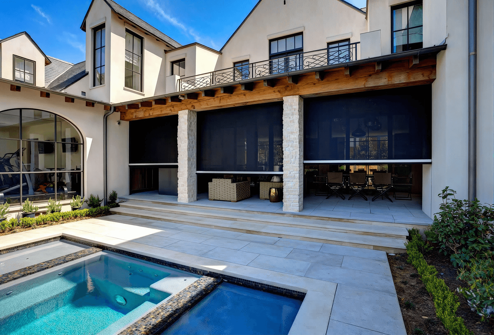Wide exterior view of a modern European-style home with a swimming pool and hot tub, featuring large dark motorized retractable shade screens installed along a covered stone-column patio.